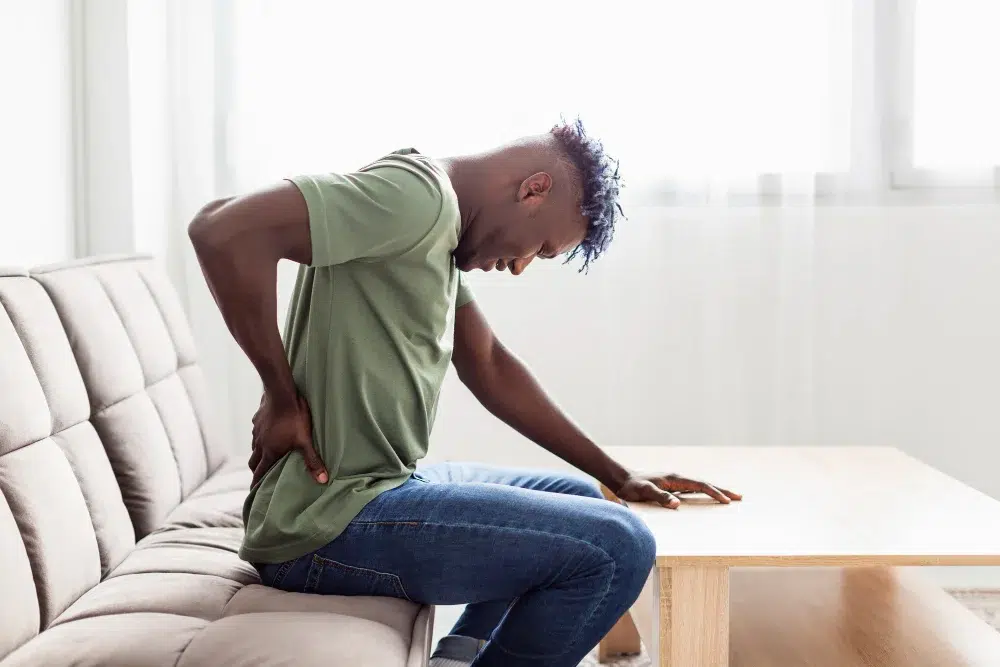 A young man with a pained expression sits on a couch, clutching his lower back in discomfort, indicating possible chronic pain issues.