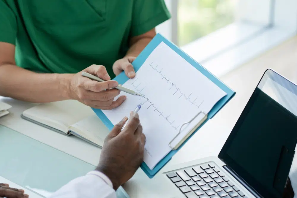 A healthcare professional in green scrubs discusses an electrocardiogram (EKG) test result with a colleague. They are pointing at the EKG printout on a clipboard while seated at a desk with a laptop and notebook.