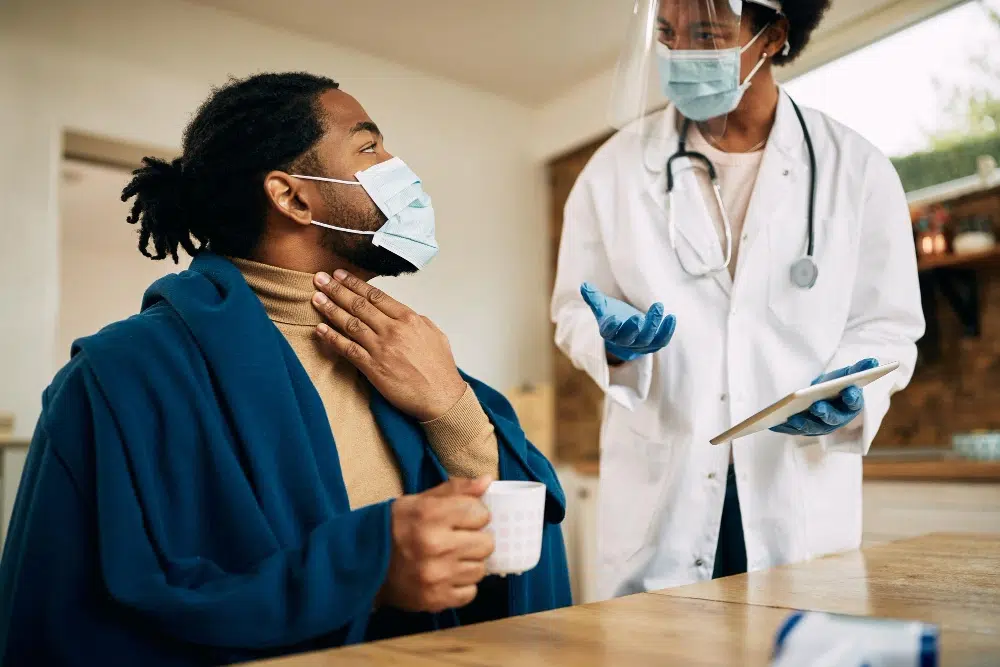 A doctor wearing a white coat, face shield, gloves, and a stethoscope is consulting a masked patient who is holding a cup and touching their throat, possibly discussing symptoms. This represents preventive care, emphasizing early intervention and health screenings to avoid serious illnesses.