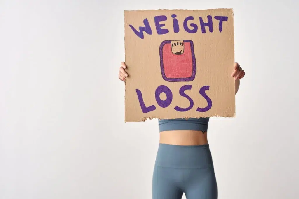 Woman in workout clothes holding a cardboard sign that says “Weight Loss” with a drawing of a scale, symbolizing a commitment to fitness and healthy living in Houston, TX