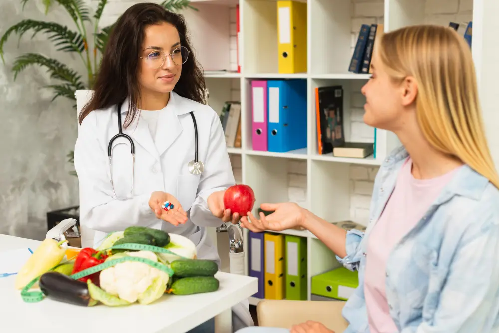 A female dietitian in a white coat and stethoscope offers a red apple in one hand and pills in the other to a young woman during a consultation. Fresh vegetables like cauliflower, cucumber, and bell peppers are on the table in front of them.