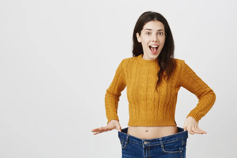 A happy and surprised young woman in a mustard sweater shows off her weight loss by wearing oversized jeans, holding the waistband out to display the difference.