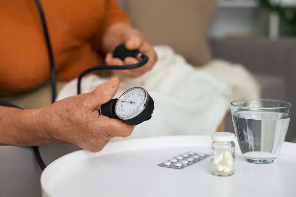 An elderly woman checking her blood pressure with medication on the table and a cup of water