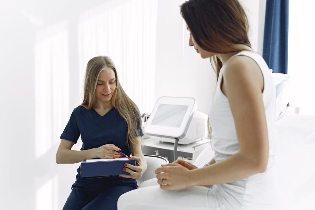 A female healthcare professional in navy scrubs talking with a patient in a white outfit inside a bright medical clinic, with medical equipment visible in the background.