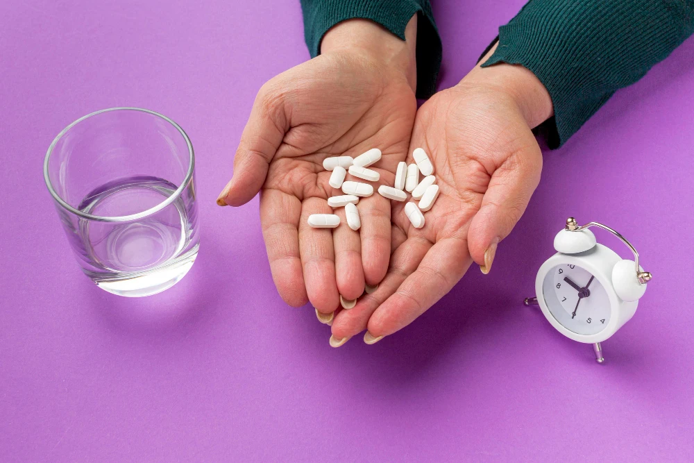 Hands holding several white pills over a purple background, with a glass of water on one side and a small alarm clock on the other, suggesting timed medication use.