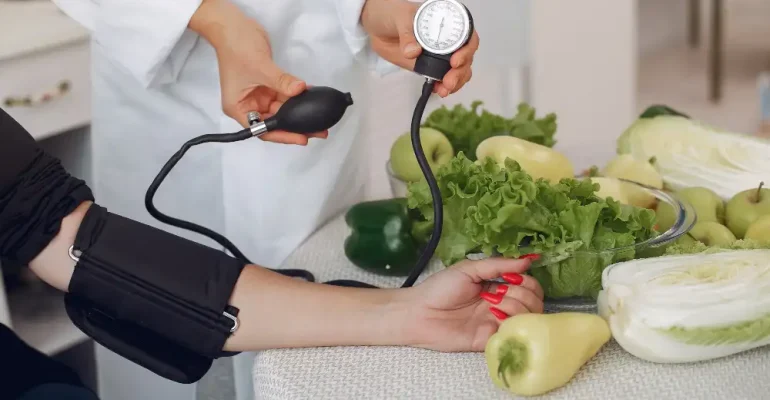 A healthcare professional measures a patient’s blood pressure using a manual cuff while fresh vegetables like lettuce, cabbage, peppers, and apples are displayed on a table, symbolizing a healthy diet.