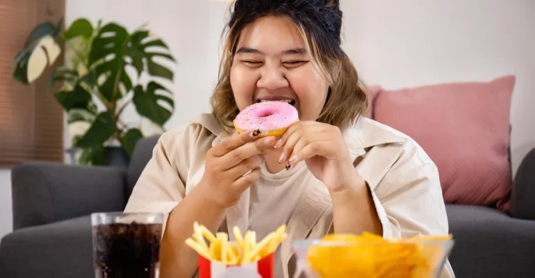 Person sitting on a couch eating a pink-frosted donut, with fast food items like fries, a burger, fried chicken, chips, and a soft drink on the table in front of them.