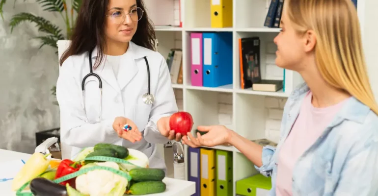 A female dietitian in a white coat and stethoscope offers a red apple in one hand and pills in the other to a young woman during a consultation. Fresh vegetables like cauliflower, cucumber, and bell peppers are on the table in front of them.