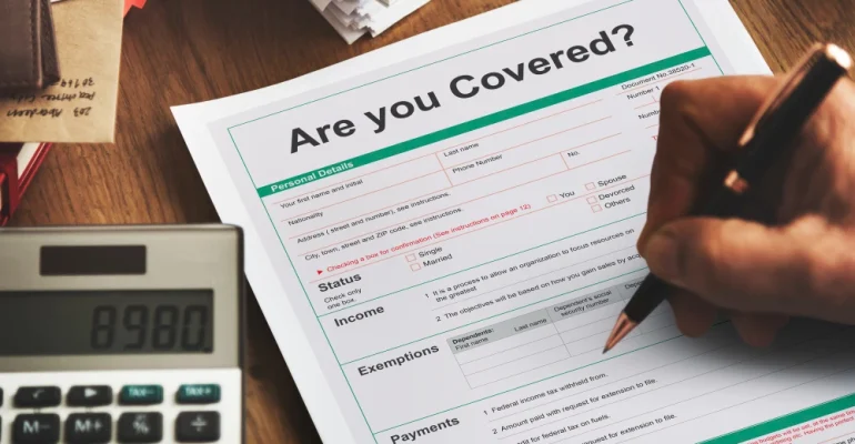 A person filling out an insurance form titled “Are you Covered?” on a desk, with a calculator and paperwork nearby