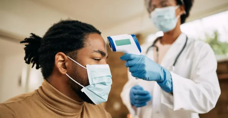 A healthcare professional wearing a face mask and gloves uses a contactless infrared thermometer to check the temperature of a patient, who is also masked. The scene illustrates preventive care measures in a medical setting.