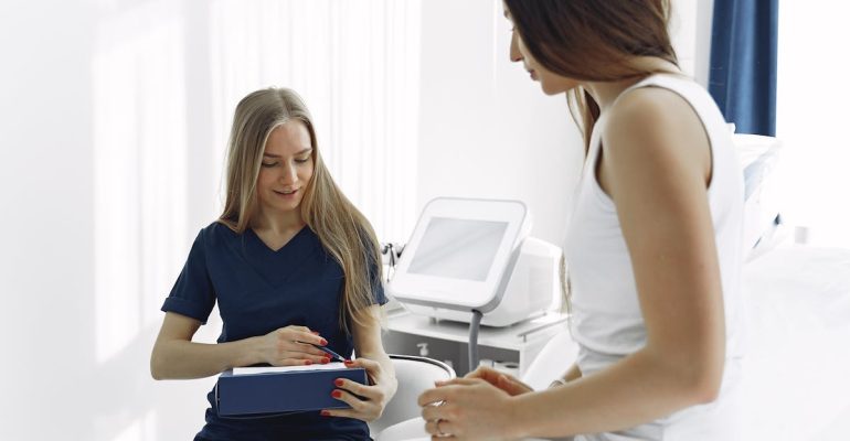 A female healthcare professional in navy scrubs talking with a patient in a white outfit inside a bright medical clinic, with medical equipment visible in the background.