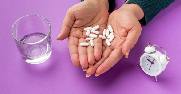 Hands holding several white pills over a purple background, with a glass of water on one side and a small alarm clock on the other, suggesting timed medication use.