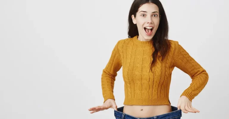 A happy and surprised young woman in a mustard sweater shows off her weight loss by wearing oversized jeans, holding the waistband out to display the difference.
